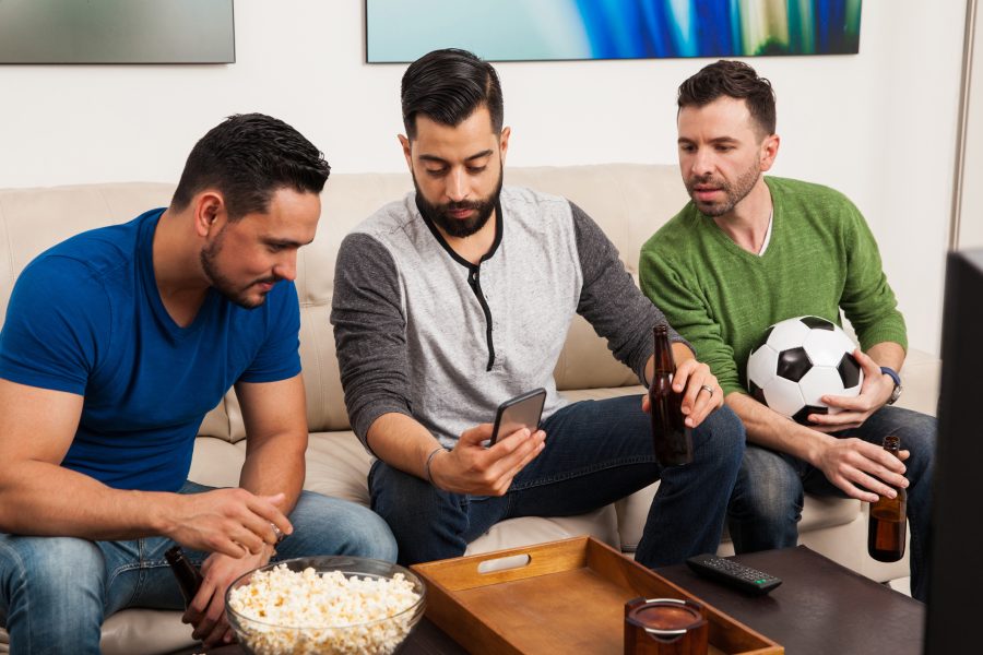 Group of friends checking their team stats on a smartphone while watching a soccer game on TV